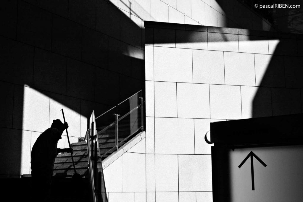 Ginza, Early Morning - The silhouette of a woman is cleaning a stair trapped inside strong shadows.