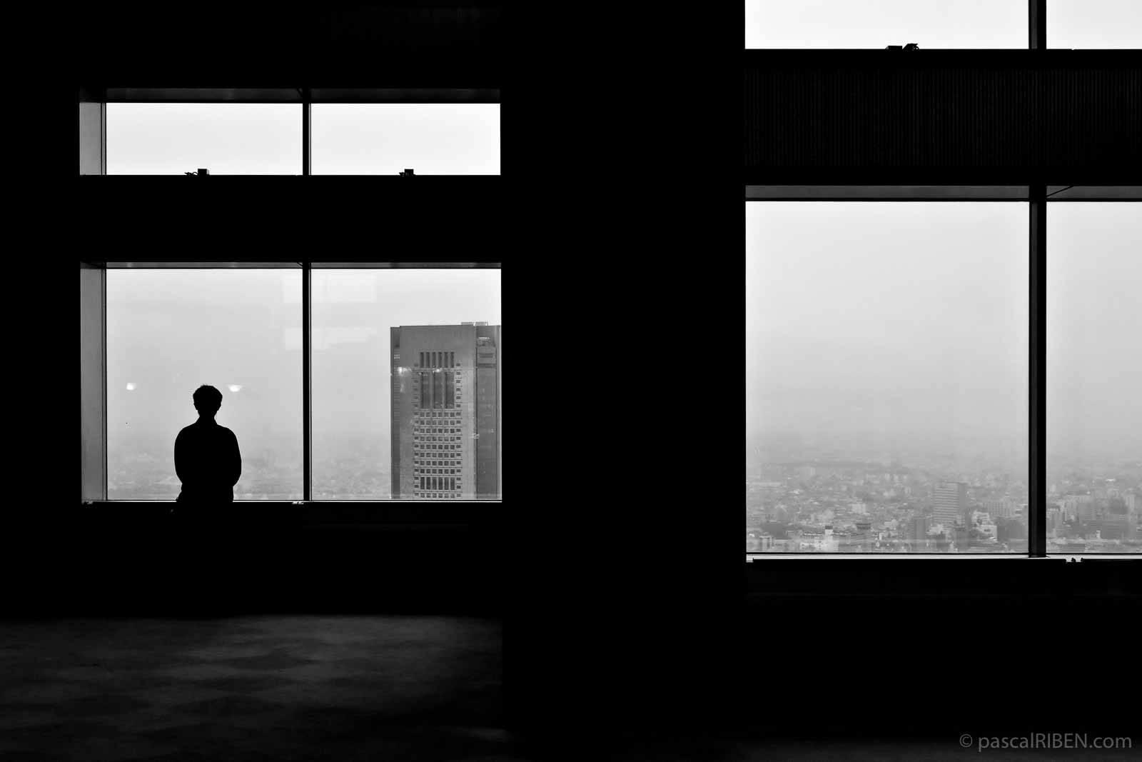 A man contemplates Tokyo across the bay window from the high floor of a building.