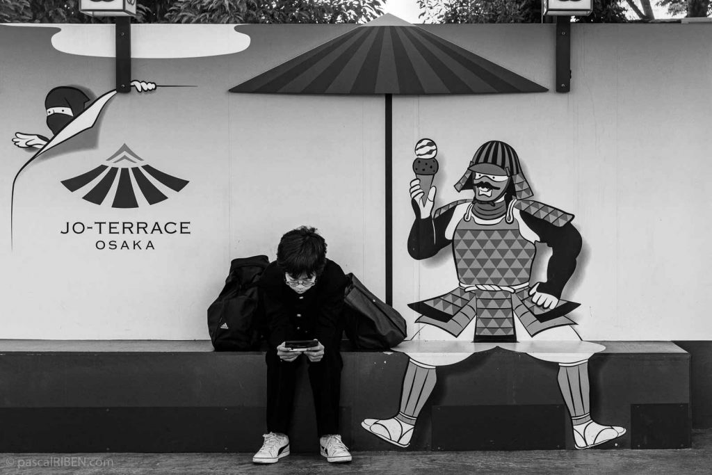 A Japanese teenager plays a video game with the real size image of a samouraï holding an ice cream behind him.