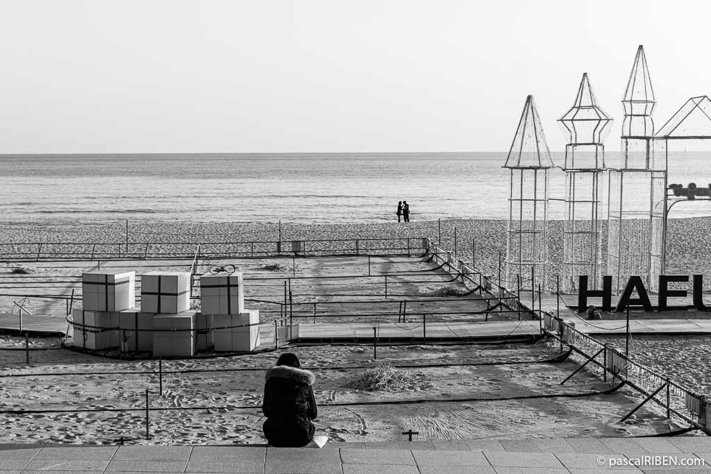 A woman, bundled up in a winter coat, sits on the steps overlooking Haeundae Beach, where remnants of Christmas installations still linger. In the near distance, the silhouette of a couple stands out against the backdrop of the sea.